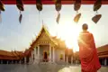 Ein Mönch in orangefarbener Robe steht im Hof eines thailändischen Tempels mit goldenem Dach, ein Programmpunkt der Besichtigung von Tempelanlagen während der Thailand-Reise.