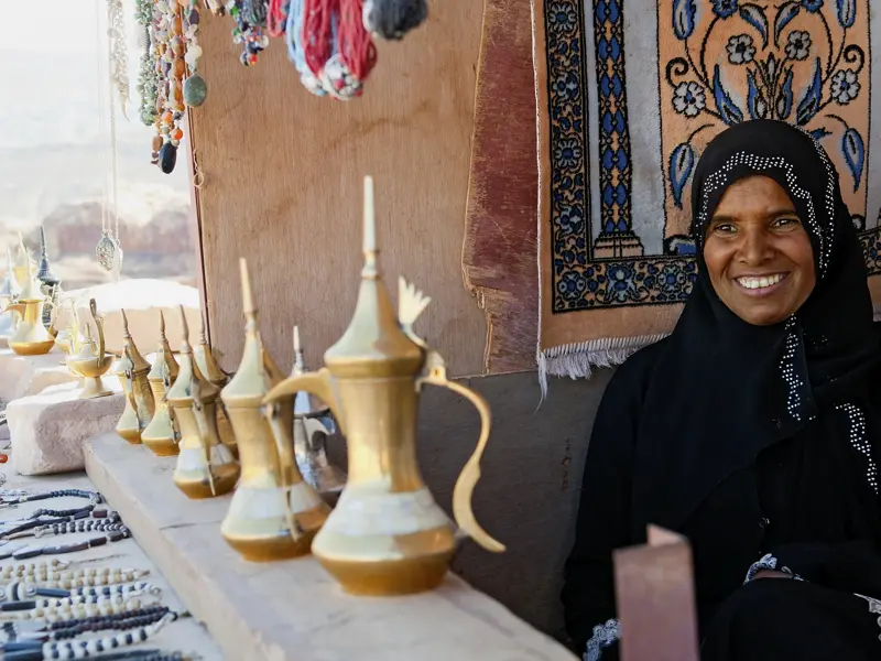 Eine  traditionell gekleidete Frau sitzt an einem Marktstand in Petra und bietet messingfarbene Kaffeekannen und Schmuck zum Verkauf an.