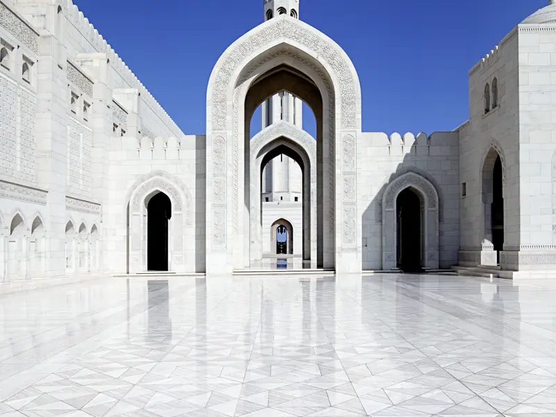 Blick auf einen der Innenhöfe der Sultan-Qaboos-Moschee in Maskat mit weiß glänzendem Marmorboden. Durch einen spitzen Torbogen erblickt man einen Teil des Minaretts.