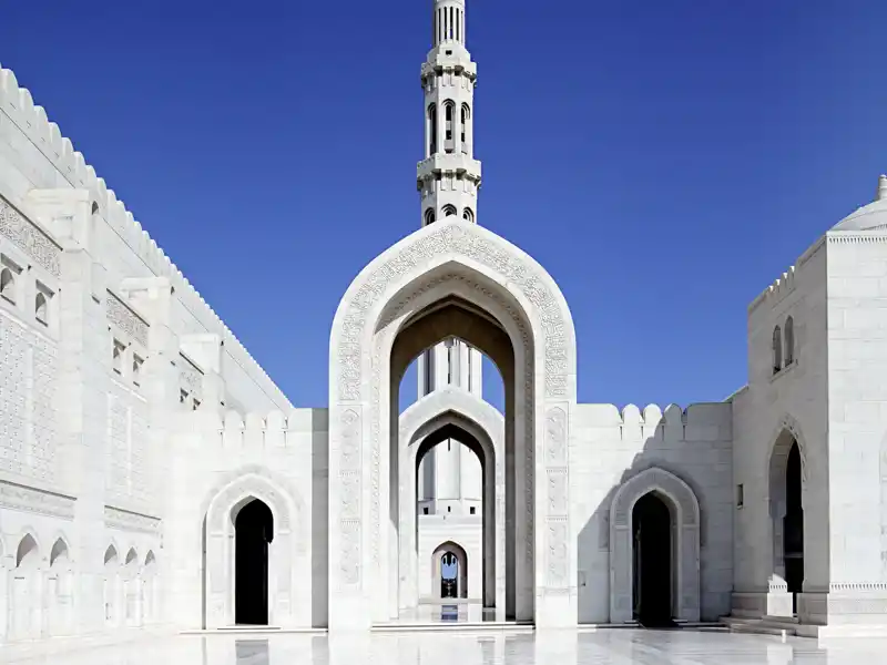 Blick auf einen der Innenhöfe der Sultan-Qaboos-Moschee in Maskat mit weiß glänzendem Marmorboden. Durch einen spitzen Torbogen erblickt man einen Teil des Minaretts.